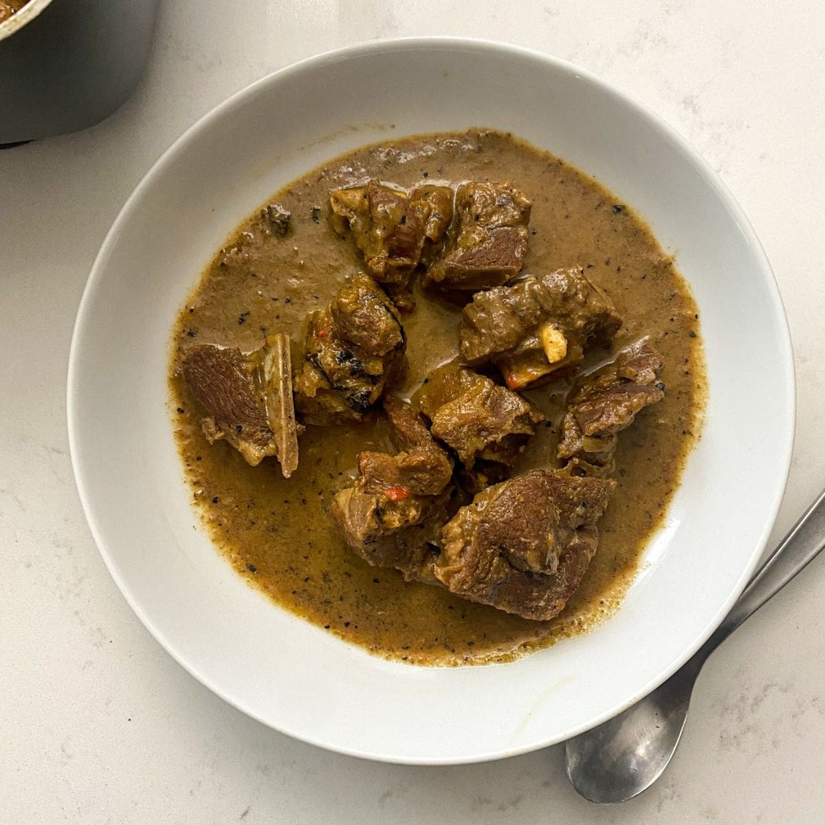 this is a picture of goat meat pepper soup in a white bowl on a marble counter top with a silver spoon